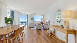 A spacious living area with a wooden dining table, two sofas, and a large window offering a view of the sea.