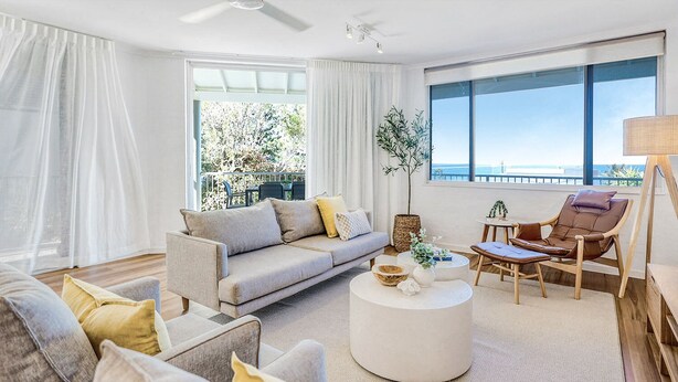 A modern living room with a grey sofa, a round coffee table, and a wooden chair. There is a large window with a view of the outdoors.