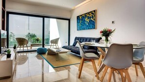 A modern living room with a sofa, dining table, and chairs. There is a large window with a view of greenery outside. - *LUXURIOUS* One Bedroom Condo behind Mamitas Beach (Gonzalo Guerrero)