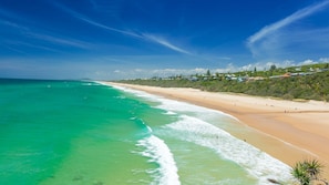 Uma praia costeira com águas turquesa cristalinas, uma faixa de areia e prédios residenciais ao fundo.