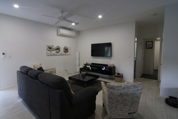 A modern living room with a dark sofa, a patterned armchair, a flat-screen TV, and a ceiling fan. - Rangimarie Holiday House (Little Mountain)