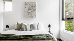 Un dormitorio con cama, almohadas, una colcha, una obra de arte enmarcada, lámparas de noche y un ventanal con vistas.