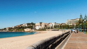 Un paseo marítimo junto a una playa arenosa, un muro de piedra y edificios al fondo.