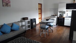Um espaço compacto para viver, com sofá, tapete estampado, pequena mesa com cadeiras, cozinha compacta e uma porta.