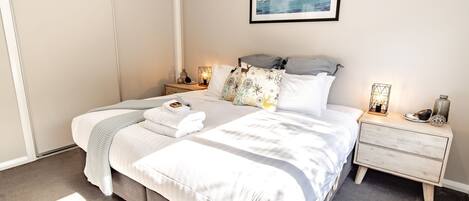 Una cama bien hecha con sábanas blancas, un edredón gris y una almohada decorativa con motivos naturales. Hay dos mesitas de noche con lámparas y objetos decorativos. Encima de la cama, en la pared, hay un cuadro con una imagen de un puerto deportivo.