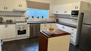 Een moderne keuken met witte kastjes, een roestvrijstalen vaatwasser, een gasfornuis en een inbouwoven. Er is een raam met zonwering, een klein eiland met een houten blad en een magnetron boven de kastjes.