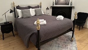 Een netjes opgemaakt bed met een donkere dekbedovertrek en decoratieve kussens, een nachtkastje met een lamp, een stoel en een open haard met daarboven een lijst met een afbeelding.
