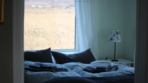 Een slaapkamer met een bed, kussens, een nachtkastje met een lamp, en een raam met uitzicht op een veld en heuvels.
