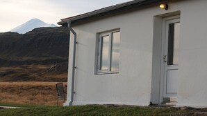 Een wit gebouw met een deur en raam, een stoel ervoor, en een berg op de achtergrond.