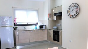 Una cucina moderna con frigorifero, microonde, piano cottura e pensili. Si vede una finestra con persiane, un orologio al muro e un vaso con fiori sul tavolo.