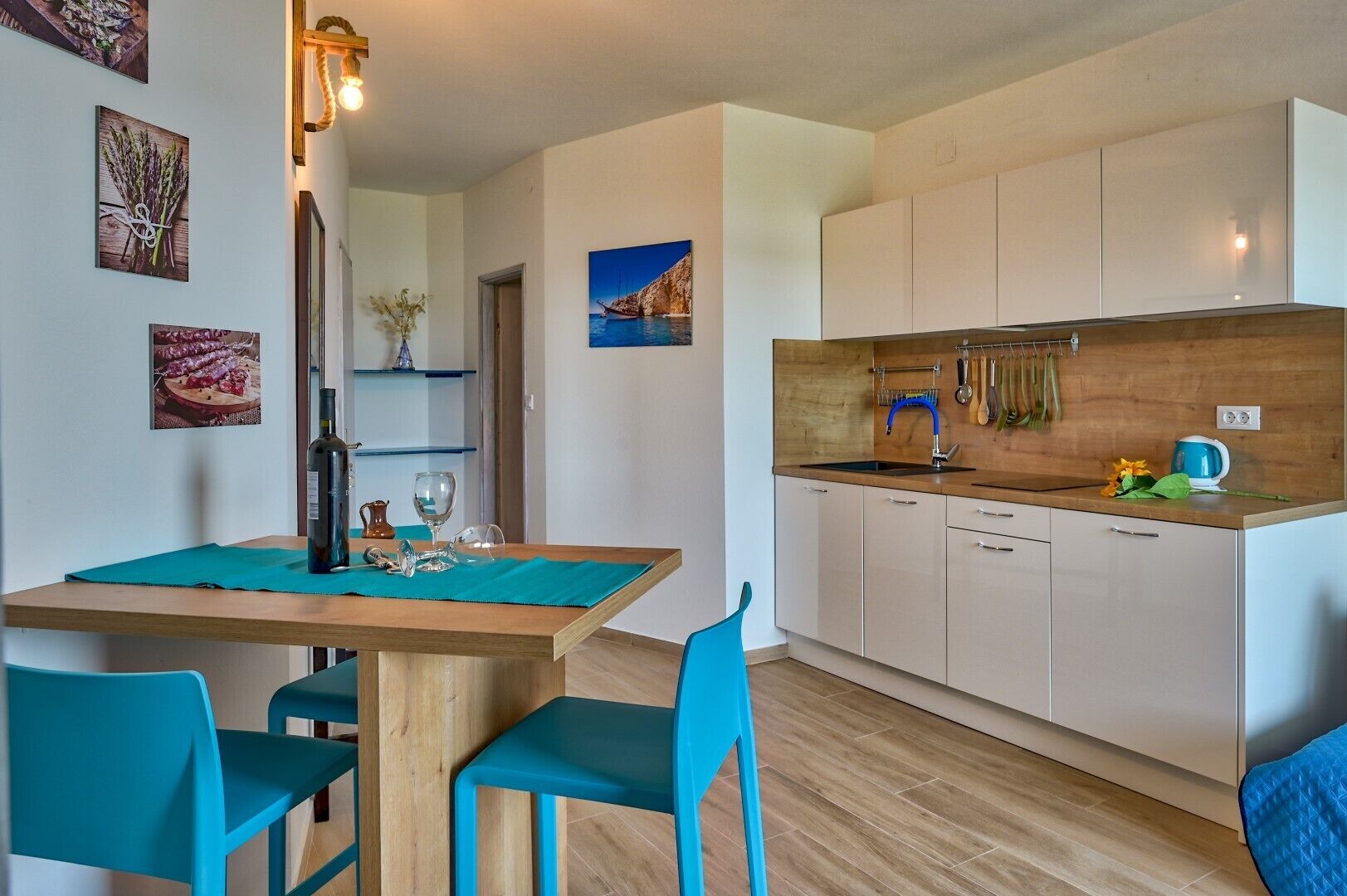 Une cuisine moderne avec des meubles blancs, un plan de travail en bois et une bouilloire bleue. On y trouve une table à manger avec des chaises bleues et un vase de fleurs.
