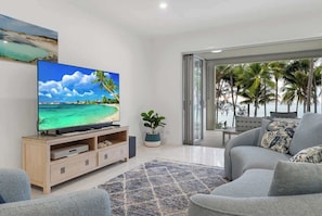A living room with a TV displaying a beach scene, a sofa, a coffee table, and a view of palm trees through a glass door.