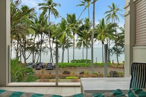 A view from a room with a large window overlooking a beach with palm trees and parked cars.