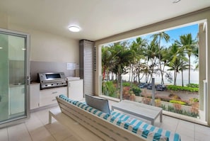 Un balcone con vista su palme e spiaggia, dotato di barbecue e area salotto con cuscini.
