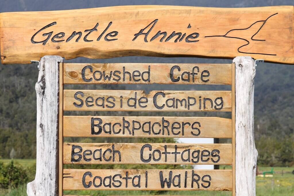 Une pancarte en bois portant le nom « Gentle Annie » et indiquant les différents services et activités proposés, tels qu’un café situé dans une ancienne étable, un terrain de camping en bord de mer, un hébergement pour les routards, des chalets de plage et des sentiers de randonnée le long de la côte.