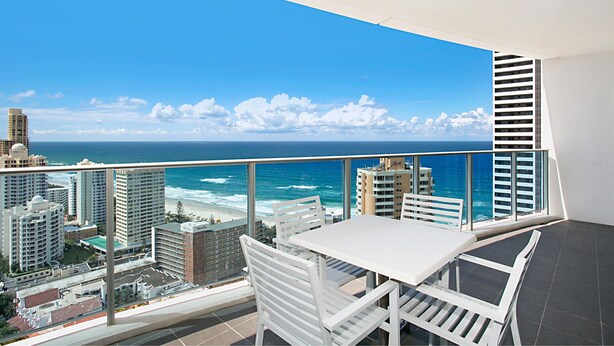 Un balcon avec une table et des chaises blanches, donnant sur une plage et l’océan, avec des bâtiments en arrière-plan.