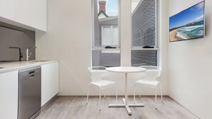 A modern kitchen with white cabinets, a dishwasher, and a window with a view of a building and a beach. There is a round table with two white chairs.