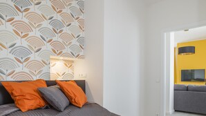 A modern bedroom with a large bed, orange and gray pillows, a patterned wall, and a view into a yellow room.
