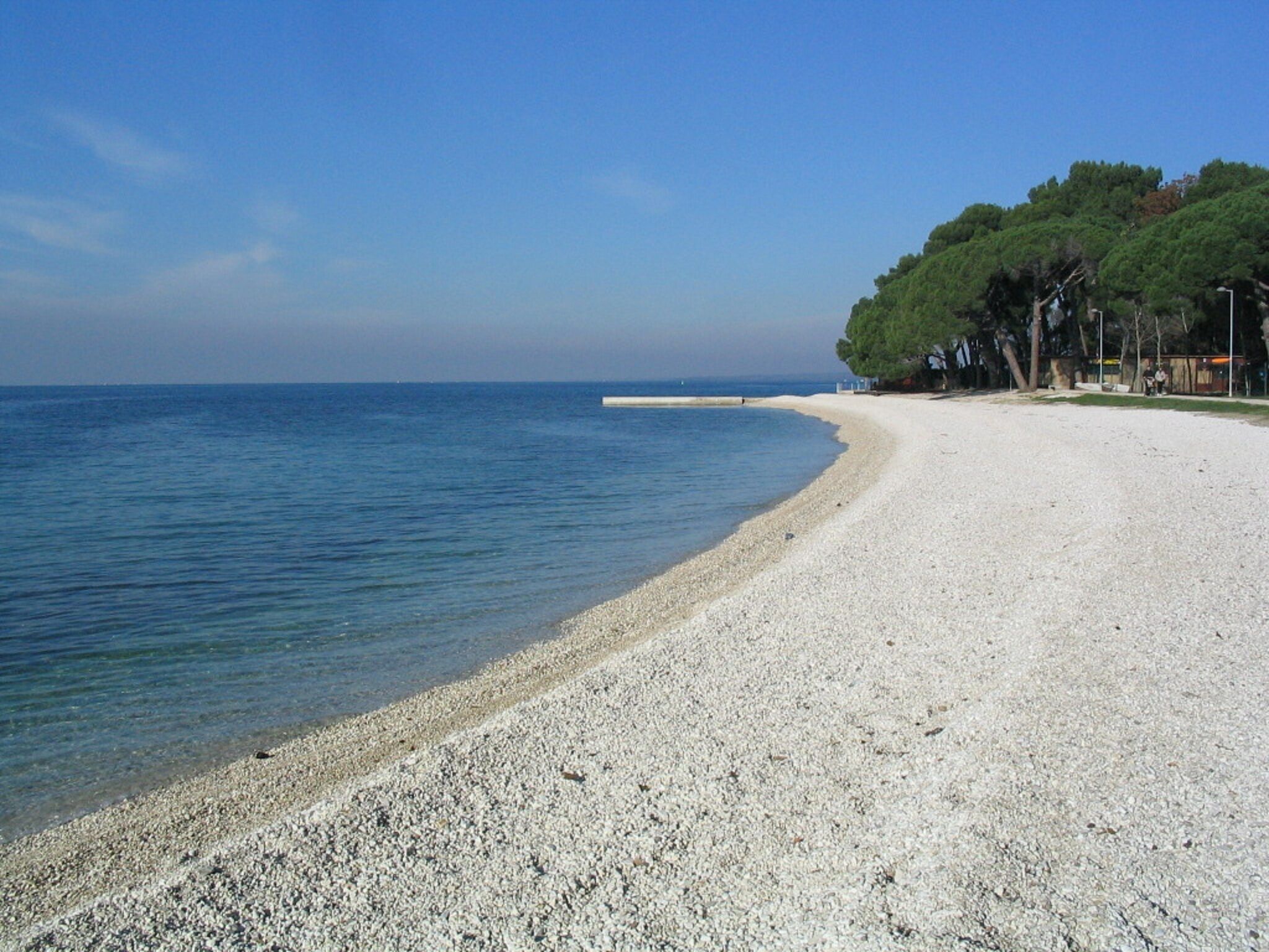 Ein Kiesstrand mit klarem, blauem Meer, einem sandigen Ufer und einer Baumreihe im Hintergrund.