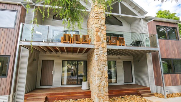 Une maison moderne à deux étages, dotée d’un pilier en pierre, d’une terrasse en bois et d’un balcon aménagé avec du mobilier d’extérieur.