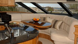 A boat interior with a kitchen area, a dining table with fruit, and a cushioned seating area.