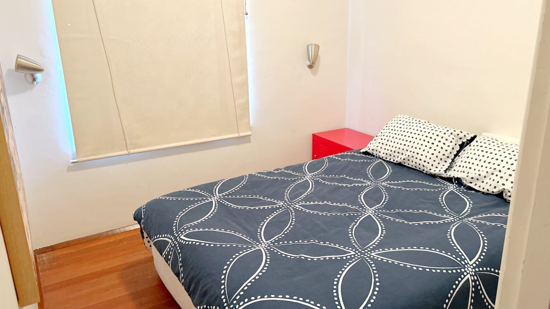 A bedroom with a bed, a red nightstand, and a window with blinds.