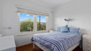 A bedroom with a bed, a bedside table, a nightstand, a window with a view of trees, and a wall-mounted lamp.