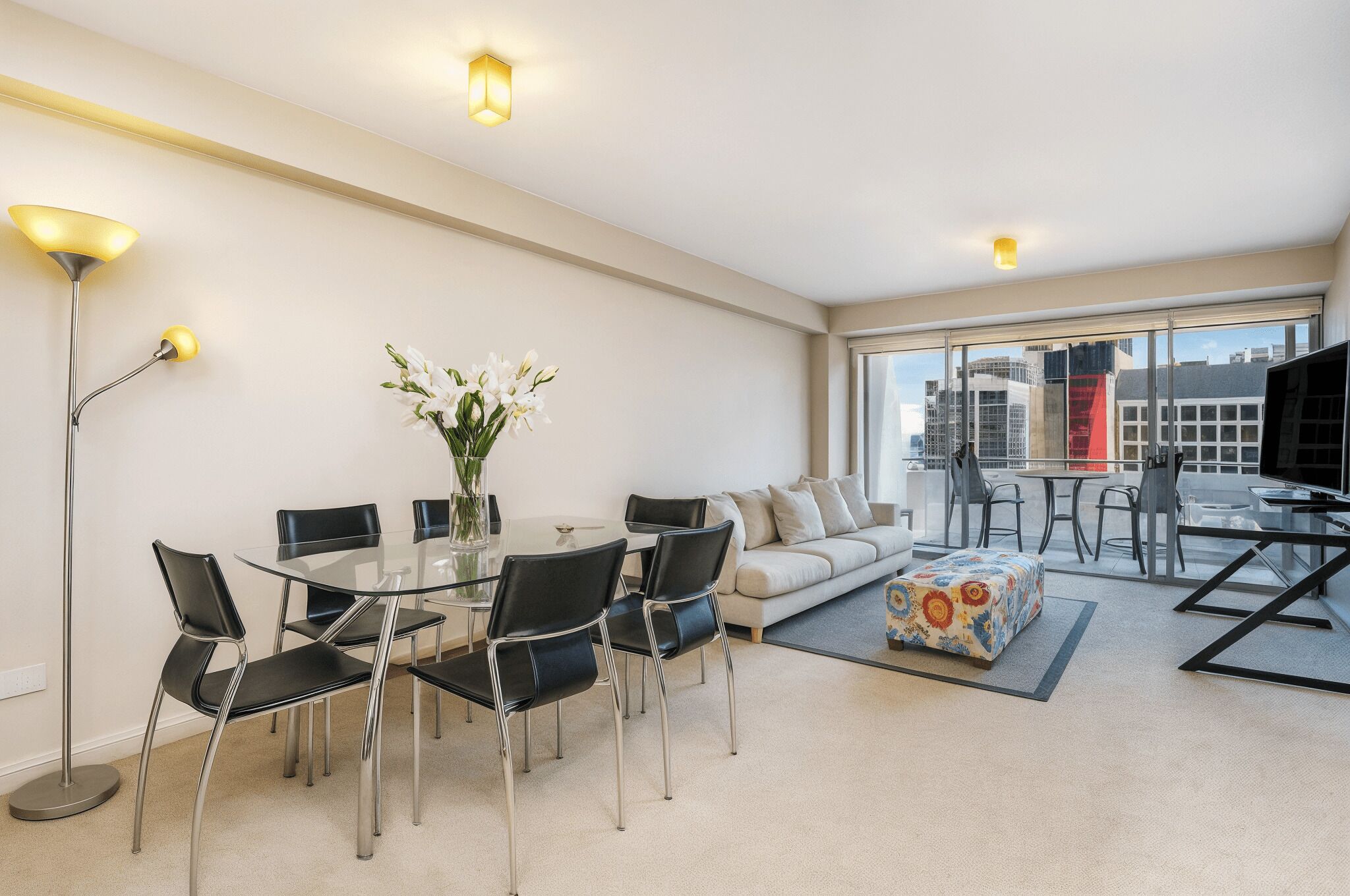 Uma sala de estar moderna com uma mesa de jantar de vidro, cadeiras pretas, sofá e televisão. Há uma janela grande com vista para a cidade.