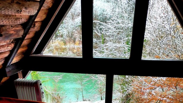 Une cabane dotée d’une grande fenêtre donnant sur un paysage enneigé. À l’intérieur, on trouve deux chaises en bois et une petite table.