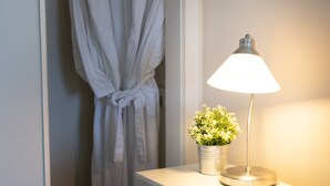 Ein Schlafzimmer mit einem Bett, einem Nachttisch mit Lampe und Topfpflanze, einer weißen Nachttischschublade und einem Kleiderschrank mit einem Bademantel darin.