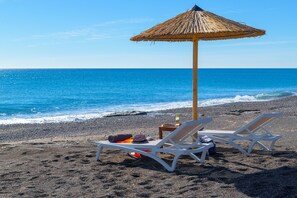 A beach with two white lounge chairs under a thatched umbrella, a small table with a bottle, and a bag on the sand.
