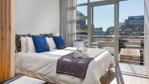 A modern hotel room with a large bed, a view of the city through floor-to-ceiling windows, and a balcony with a railing. - Backup-Powered City Apartment with Breathtaking Views on Popular Bree Street  (Cape Town)