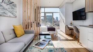 A modern living room with a sofa, coffee table, and a view of the city through large windows. - Backup-Powered City Apartment with Breathtaking Views on Popular Bree Street  (Cape Town)