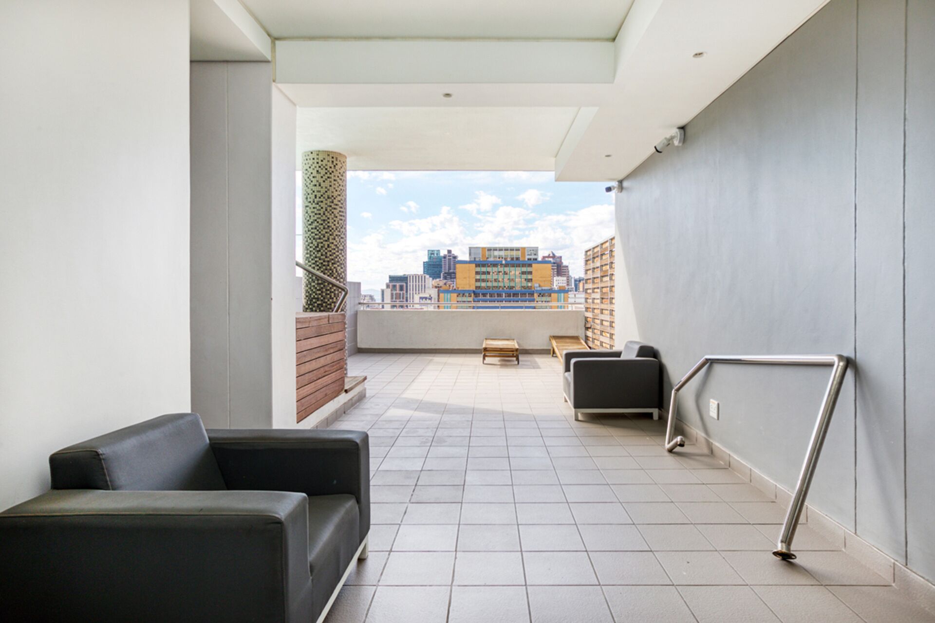 A modern rooftop terrace with a view of a cityscape, featuring a seating area with a sofa and a table, and a metal railing.