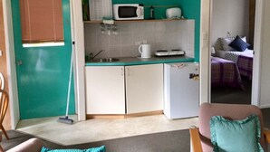 A compact kitchen with a microwave, toaster, and small refrigerator. There is a window with blinds and a door leading to a bedroom with a bed and a nightstand.