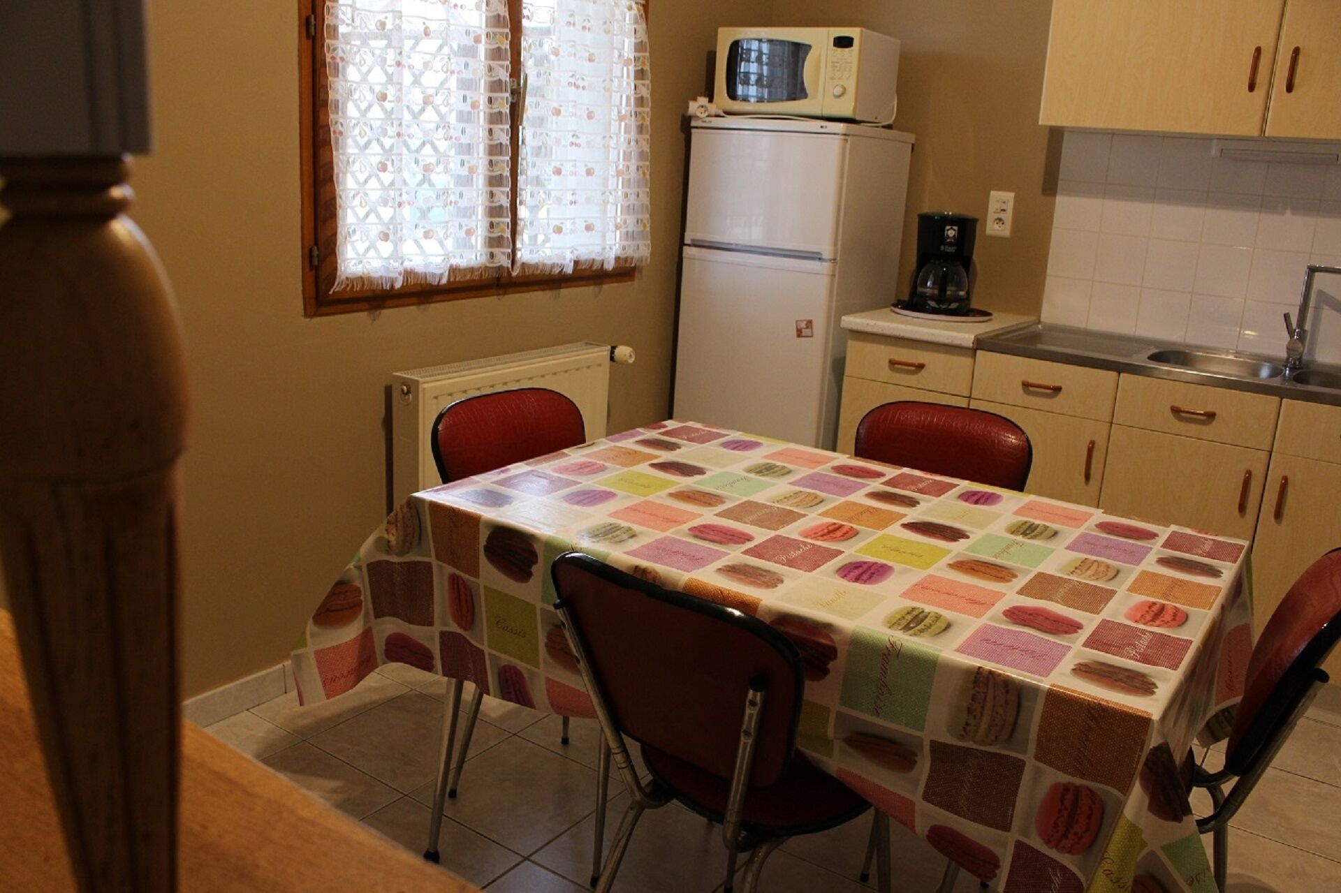 A compact kitchen with a refrigerator, microwave, and coffee maker. There is a table with a colorful tablecloth and two red chairs.