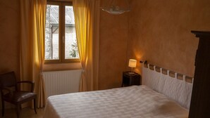 A bedroom with a bed, a chair, a nightstand with a lamp, a window with curtains, and a radiator. - Countryside house - Le Rochepot - Cote d'Or (La Rochepot)