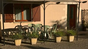 A covered outdoor seating area with tables and chairs, potted plants, and a building with windows and a door in the background. - Countryside house - Le Rochepot - Cote d'Or (La Rochepot)