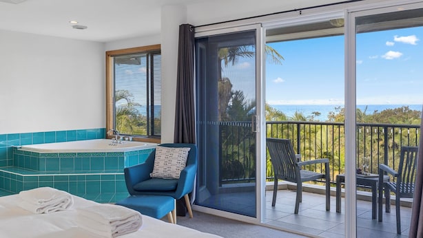 Una habitación con cama, una silla azul, bañera y un balcón con sillas y vistas al océano.