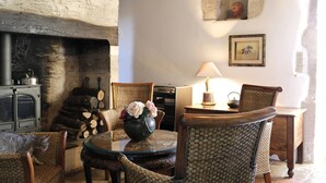Una habitación acogedora con chimenea de piedra, muebles de mimbre, una mesa redonda de cristal con un florero y una lámpara sobre una mesita de madera.