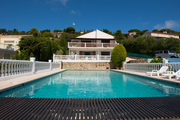 Une piscine avec une terrasse en bois au premier plan, une clôture blanche et un bâtiment à deux étages doté d’un balcon en arrière-plan.