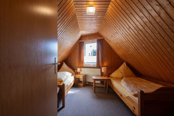 Ein gemütliches Schlafzimmer im Dachgeschoss mit Holzwänden und schrägen Decken. Es beinhaltet zwei Betten, einen kleinen Tisch und ein Fenster mit Blick ins Freie.