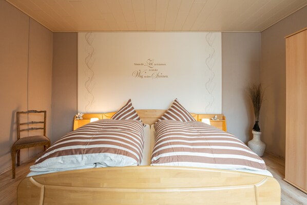 A bedroom with a wooden bed, striped pillows, a nightstand, a chair, and a decorative wall with text.