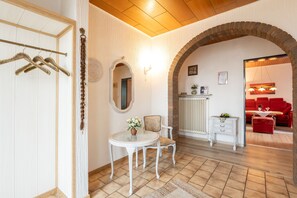 A room with a white door, a coat rack, a round table with a vase, a chair, a white dresser, a red sofa, and a brick archway.