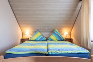A bedroom with a wooden ceiling, two bedside lamps, a bed with a blue and yellow striped bedspread, and matching pillows. - Apartment Helene EG 35179 - Apartments Engelmann 35107 OG and Helene 35179 EC (Holtland)