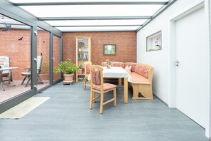 A sunlit patio with a brick wall, a glass door, a table with chairs, a bench, and a bookshelf. - Apartment Helene EG 35179 - Apartments Engelmann 35107 OG and Helene 35179 EC (Holtland)