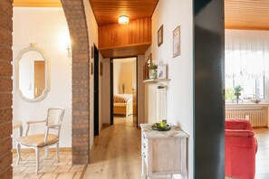 A hallway with a wooden floor, a white chair, a small table with a vase, and a red chair in the background.