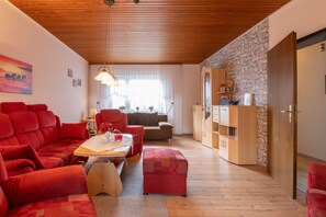 A cozy living room with red furniture, a wooden ceiling, a stone wall, and a dining area with natural light. - Apartment Helene EG 35179 - Apartments Engelmann 35107 OG and Helene 35179 EC (Holtland)