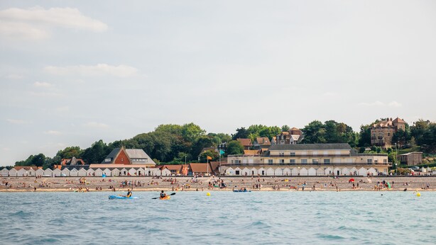Une zone côtière bordée d’une rangée de cabanes de plage le long du rivage, un grand bâtiment avec un toit rouge et, au loin, une structure qui ressemble à un château.