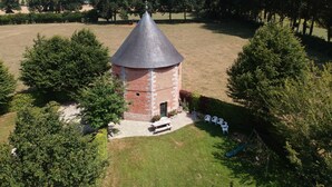 Une petite tour en briques, surmontée d’un toit conique, est entourée d’une zone herbeuse et d’arbres.
